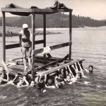 Pinewood swim raft at Sly Park, summer of 1962. Larry Slater on the far left, Ron Parker on the far right, Margaret Slater on the raft. Students unidentified.